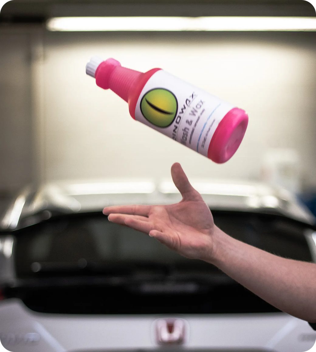 A hand tossing a pink bottle labeled 'Wash & Wax' in front of a car.